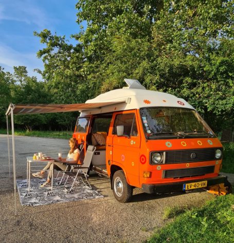 Oranje camper met luifel, man en vrouw ontspannen op stoelen in de natuur.